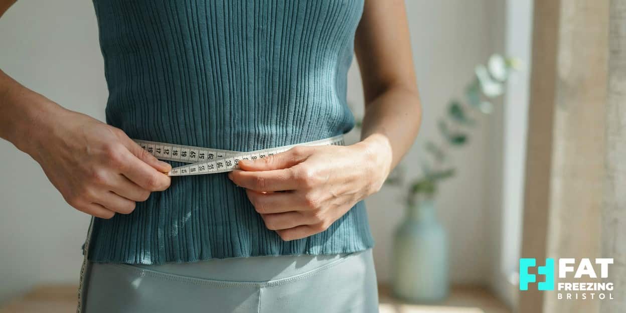 A person’s hands holding a cloth tape measure around their waist over a teal top, taking an at-home waist measurement in soft daylight — a simple alternative to BMI we encourage at our fat freezing clinic in Bristol