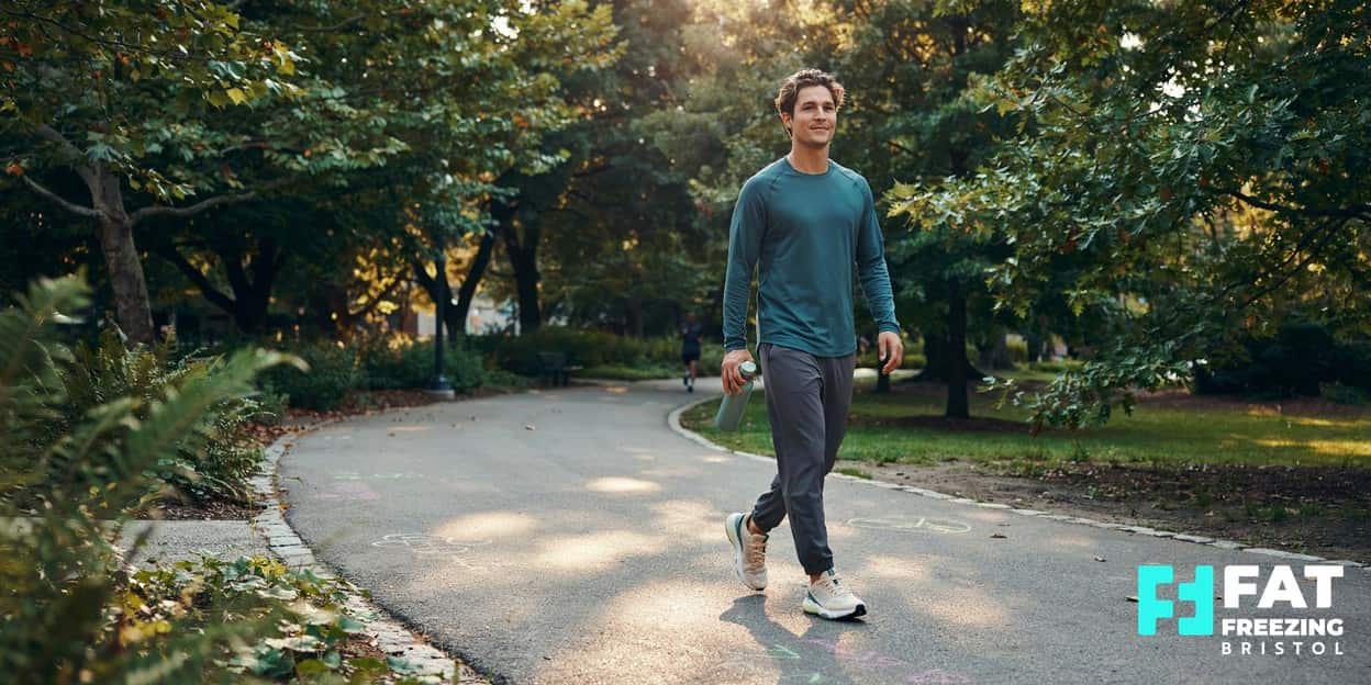 A person taking a relaxed walk along a leafy park path with a water bottle in soft morning light, illustrating the gentle movement encouraged during fat freezing aftercare recovery