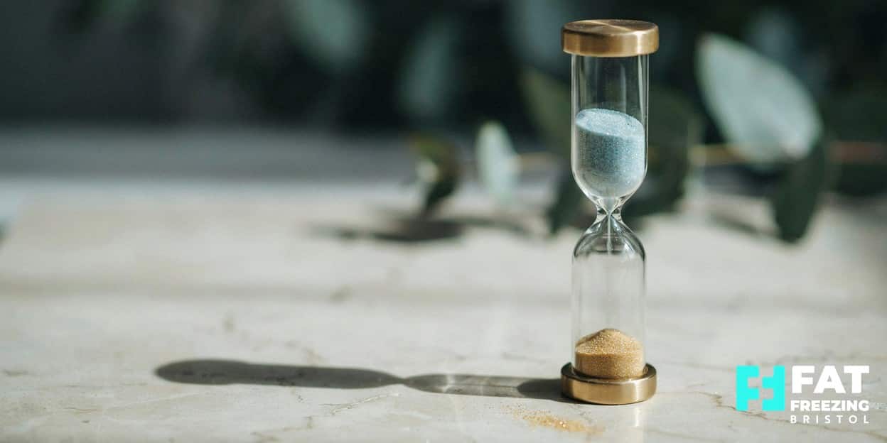 Still-life of a glass hourglass with frosted blue sand above and warm gold sand below in soft daylight, representing the patient weeks-long timeline for fat freezing results to emerge
