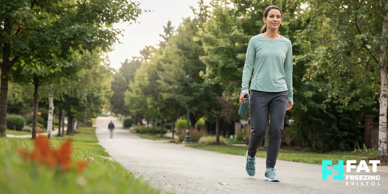 An adult in activewear walking with a relaxed stride along a tree-lined path in soft morning light, carrying a water bottle — suggesting the sustainable habits that complement fat freezing results