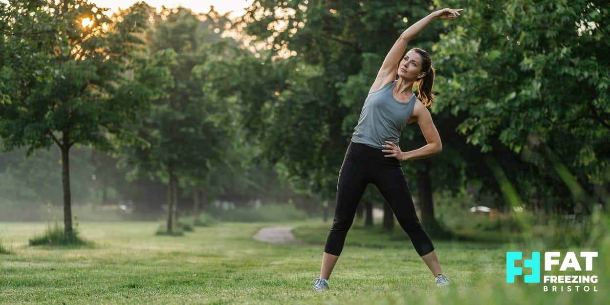 A woman in activewear doing a standing side-stretch outdoors at sunrise in a park, reflecting the active lifestyle that complements fat freezing treatment