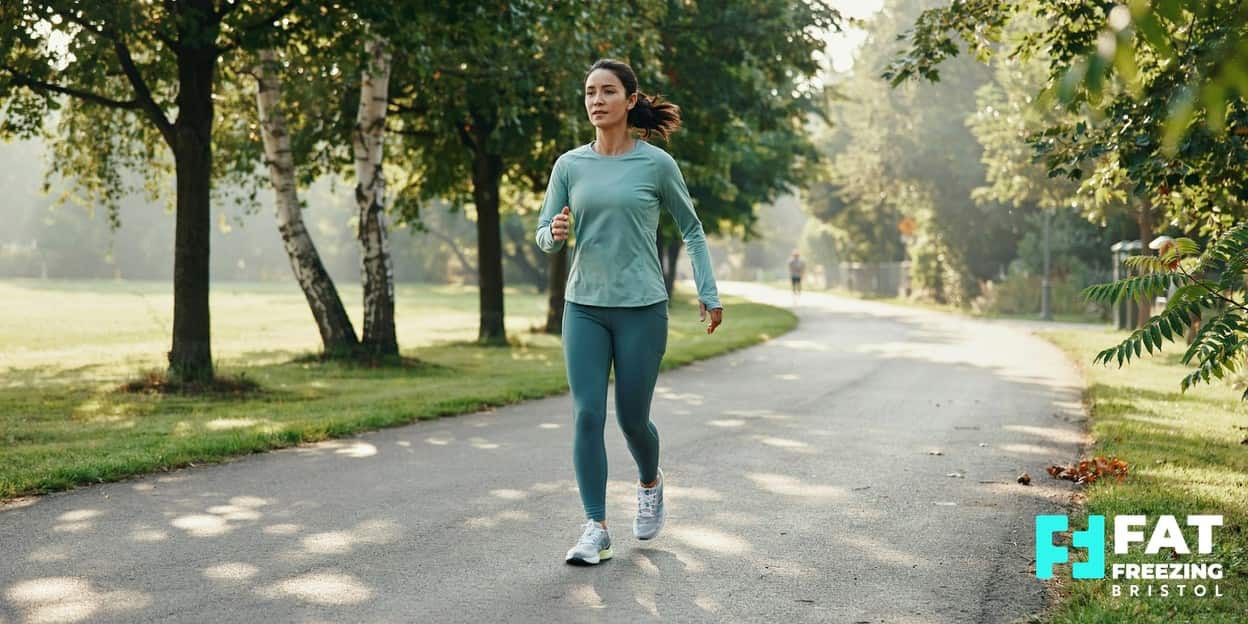 An adult in activewear walking briskly along a sunlit tree-lined path in soft dappled morning light, depicting the healthy-lifestyle context of weight management alongside body-contouring options like fat freezing in Bristol
