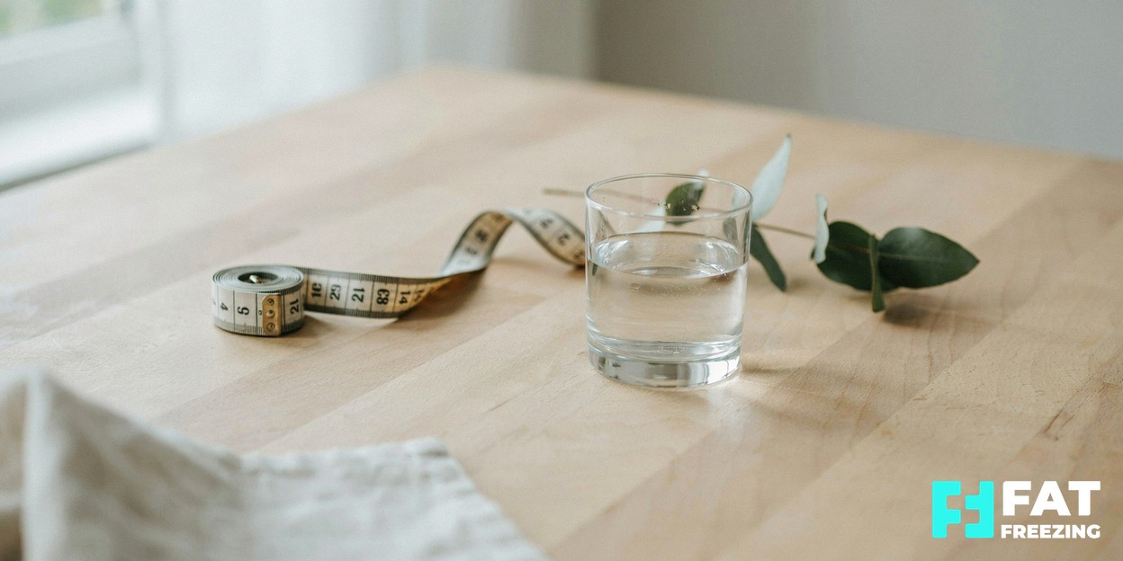 A soft-focus close-up of a cloth tape measure resting on a clean light wooden surface beside a glass of water in calm daylight