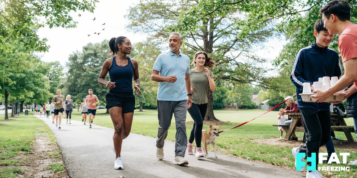 A diverse group of adults of mixed ages walking and jogging together at a friendly outdoor community 5K event in a green park on a bright morning